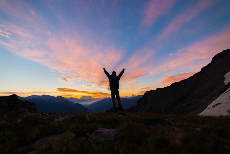 Man standing on mountain top outstretching arms, sunrise light colorful ...